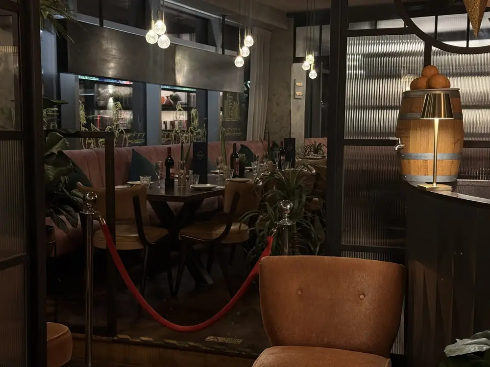Tables and banquettes at The Reach Riverside restaurant in Fulham Reach, with a brass lamp glowing on a barrel