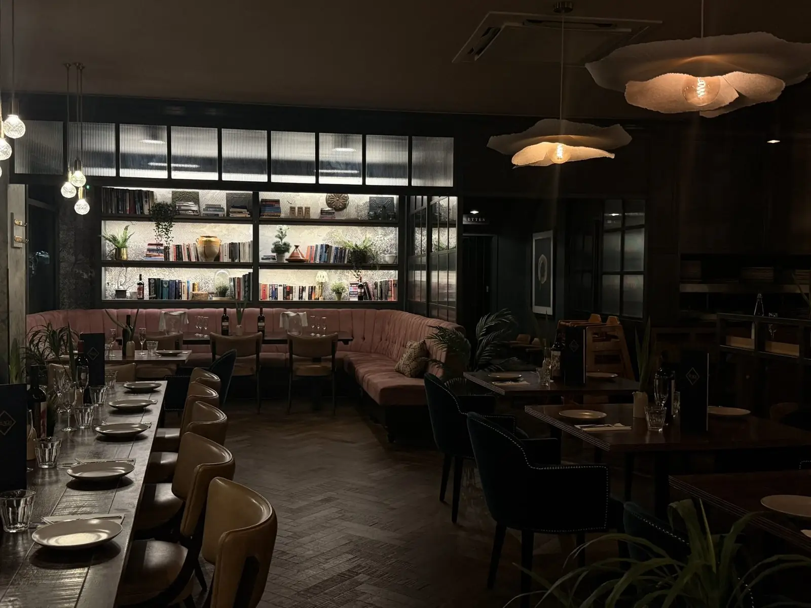 A long table laid for a group dinner beneath wall-lit bookshelves at The Reach Riverside, Fulham Reach
